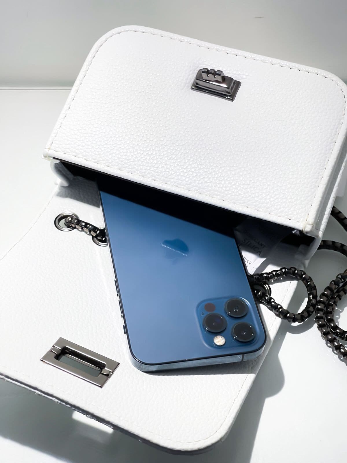 Celular azul sendo inserido na bolsa branca matelassê, aberta, para demonstrar o tamanho e a capacidade interna.