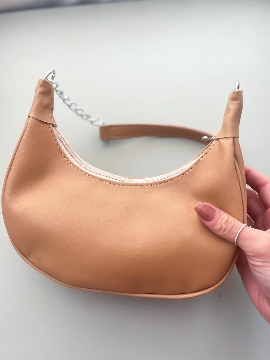 Mão segura bolsa de ombro feminina bege clara (nude) em formato meia-lua, com zíper branco e corrente prateada