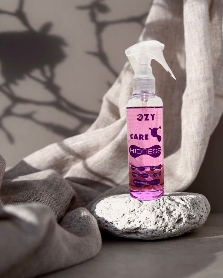 Frasco de spray Ozy Care Hidress Over Care com líquido roxo sobre uma pedra, com fundo de tecido texturizado e sombra.