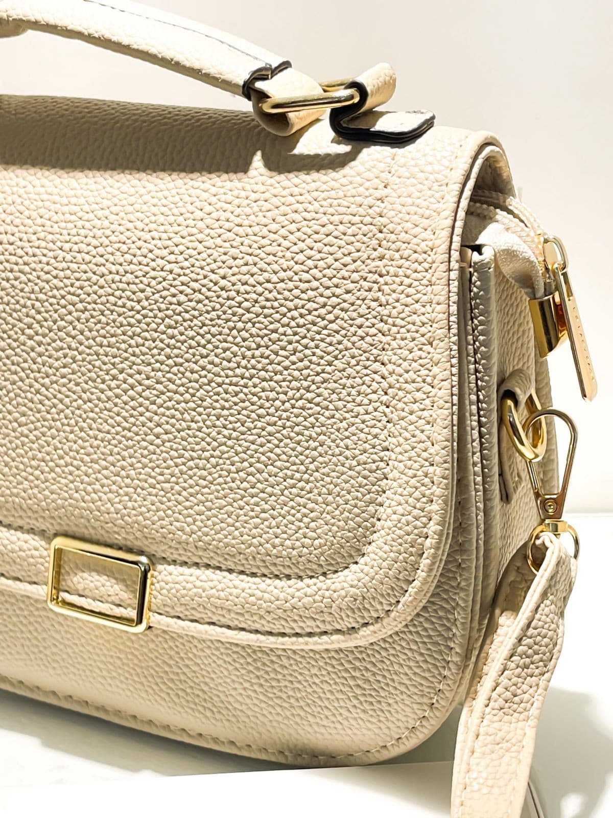 Close-up da lateral de bolsa bege texturizada, destacando fecho dourado, zíper lateral e gancho da alça.