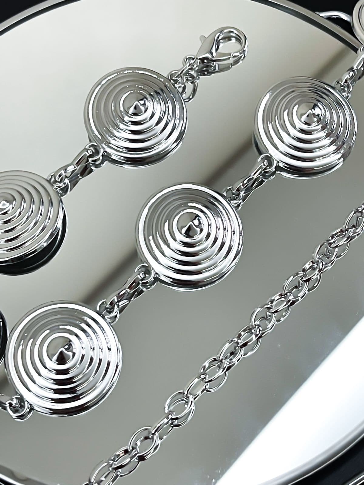 Cinto de corrente prateada com discos redondos interligados com relevos em espiral, refletindo em uma superfície clara.