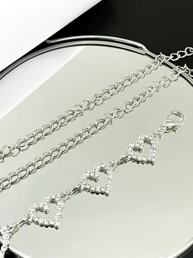 Cinto de corrente prateada com três corações cravejados de strass, refletindo em um fundo espelhado.
